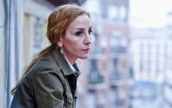 A woman with reddish-brown hair in a ponytail and wearing a green jacket leans on a balcony railing, looking thoughtfully into the distance, with blurred buildings in the background.