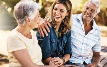 A young woman sits outdoors on a bench, smiling and holding hands with an older woman, while an older man with his arm around the woman looks on, all appearing happy and relaxed together.