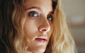 Close-up of a young woman with long, wavy blonde hair and blue eyes, looking slightly to the side with a neutral expression. The background is softly blurred.