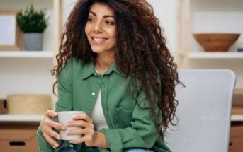 A young woman with long, curly hair sits on a chair, smiling and holding a mug. She is wearing a green shirt and jeans, with shelves and decorative items in the background.