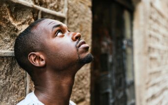 A young man with short hair and a trimmed beard stands next to a stone wall, looking upwards with a thoughtful expression. The background is slightly blurred, highlighting the texture of the wall.