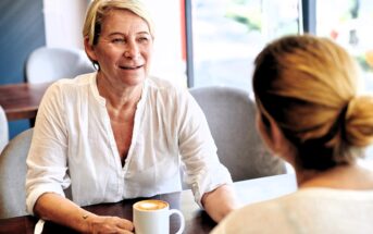 Two women sit across from each other at a cafe table, talking. One woman, with short blonde hair and a white shirt, holds a coffee mug and smiles warmly at the other, who has her back to the camera.