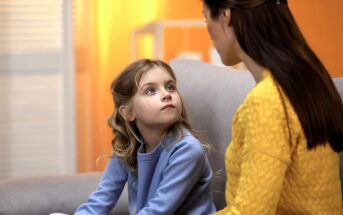 A young girl with blonde hair, wearing a light blue sweater, sits on a couch looking up attentively at a woman in a yellow sweater. The background is softly lit with warm yellow tones.