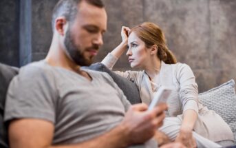 A woman sits on a couch looking concerned at a man who is focused on his phone, ignoring her. The atmosphere appears tense and distant between the two.