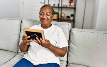An older woman sits on a light-colored sofa, smiling warmly as she looks at a framed photo she holds in her hands. Shelving with plants and decor is visible in the softly lit background.