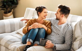 A woman and a man sit closely together on a striped couch, smiling and looking at each other. The woman wears a brown sweater and jeans, while the man holds a mug and wears a striped shirt. A plant is visible in the background.
