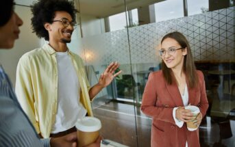 Three people stand indoors holding takeaway coffee cups, engaged in conversation. One man gestures while smiling, and a woman in a rust blazer listens and smiles. They are near a glass wall with a geometric pattern.