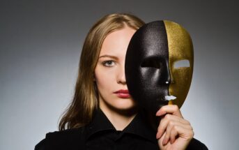 A woman with long blond hair holds a black and gold half-mask in front of her face, covering one eye, while the other half of her face is visible. She has a serious expression and stands against a gray background.