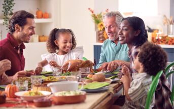 A diverse family of five joyfully shares a Thanksgiving meal at a decorated dining table, featuring a roast turkey, vegetables, and autumn-themed decor in a bright kitchen.