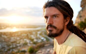 A man with long brown dreadlocks and a beard gazes thoughtfully into the distance, sitting outdoors with a blurred view of a sunlit landscape, including water and hills, in the background.