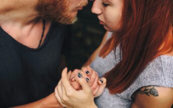 A close-up of a couple embracing tenderly, holding hands and touching foreheads. The woman has red hair and a tattoo on her arm; the man has a short beard. Their eyes are closed, conveying intimacy and affection.