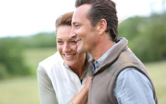 A smiling couple stands close together outdoors, with the woman resting her hand on the man's shoulder. They both appear happy and are dressed in casual outdoor clothing, with a blurred green landscape in the background.