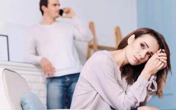 A woman sits on a couch looking upset, resting her head on her hands. In the background, a man stands and talks on a phone, appearing indifferent or distracted. The scene suggests tension or emotional distance.
