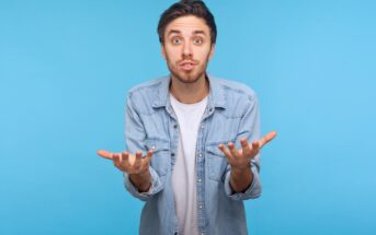 A young man in a light denim shirt and white t-shirt stands against a blue background, looking confused or questioning, with both hands raised and palms facing up.