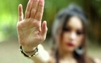 A person with long hair holds their hand up toward the camera, palm facing forward, signaling “stop.” The background is blurred and green, and the person’s face is out of focus.