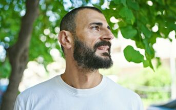 A bearded man in a white t-shirt stands outdoors, looking to his right. Green leaves and tree branches create a natural background, with sunlight filtering through.