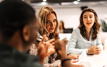 Three people sit at a table in a meeting. A woman with blonde hair in the center looks directly at the camera while listening to a man in the foreground. Another woman in the background is talking and holding papers.