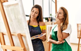 Two women stand by an easel in a bright art studio. One holds a palette and brush while painting, and the other, wearing an apron, watches thoughtfully with her hand on her chin.