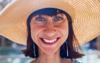 A woman with short brown hair smiles brightly, wearing a large, wide-brimmed straw hat. Sunlight highlights her face and blue eyes. The background is softly blurred, suggesting an outdoor setting.