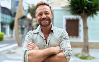 A smiling man with a beard and short hair stands outdoors with his arms crossed, wearing a light striped shirt. The background features buildings, trees, and a street, creating a relaxed urban setting.