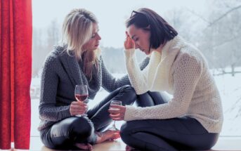 Two women in cozy sweaters sit by a window holding wine glasses. One woman appears upset with her head in her hand, while the other gently comforts her. Snow is visible outside the window.