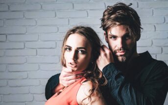 A woman in an orange top stands in front of a man in a black shirt. The man is gripping her chin and neck with one hand. Both have serious expressions and stand against a white brick wall.