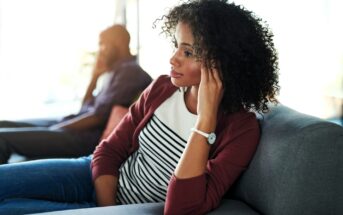 A woman with curly hair sits thoughtfully on a couch, looking away, with her hand touching her ear. A man in the background sits on another part of the couch, slightly out of focus.