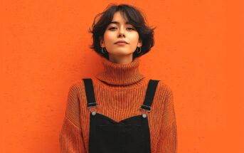 A young person with short dark hair, wearing hoop earrings, a ribbed orange turtleneck sweater, and black overalls stands against an orange textured background, looking confidently at the camera.