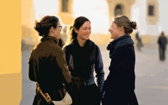 Three women stand outdoors in conversation, smiling at each other. They wear dark coats and have their hair tied back. The background is softly blurred, suggesting a bright, sunlit day in an urban setting.