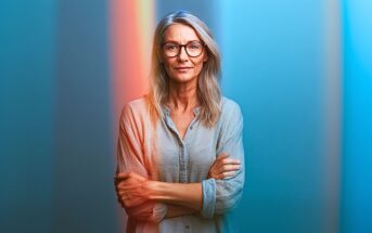 A confident middle-aged woman with long blond hair and glasses stands with arms crossed, wearing a light blue shirt, against a blue background with vertical colored light streaks.