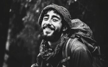 A young man with a beard and curly hair smiles warmly at the camera, wearing a scarf and a backpack. The black-and-white photo gives a feeling of adventure and outdoor exploration.