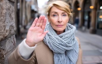 A middle-aged woman with blonde hair stands outdoors in a city street, wearing a beige coat and a chunky gray scarf. She holds up her hand, palm forward, with a serious expression, as if signaling stop.