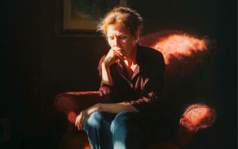 A woman sits on a red armchair in a dimly lit room, resting her chin on her hand and looking down with a thoughtful or somber expression. Warm light highlights her face and the chair.