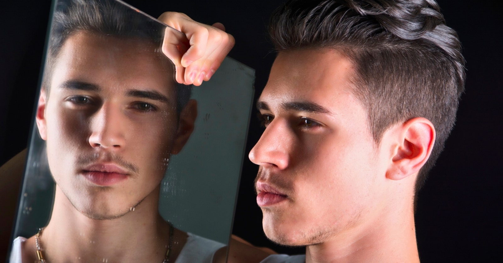 A young man with dark hair looks into a mirror, his reflection showing a pensive expression. He holds the mirror up with his left hand, which is slightly visible. The background is dark, emphasizing the contrast between the man and his reflection.