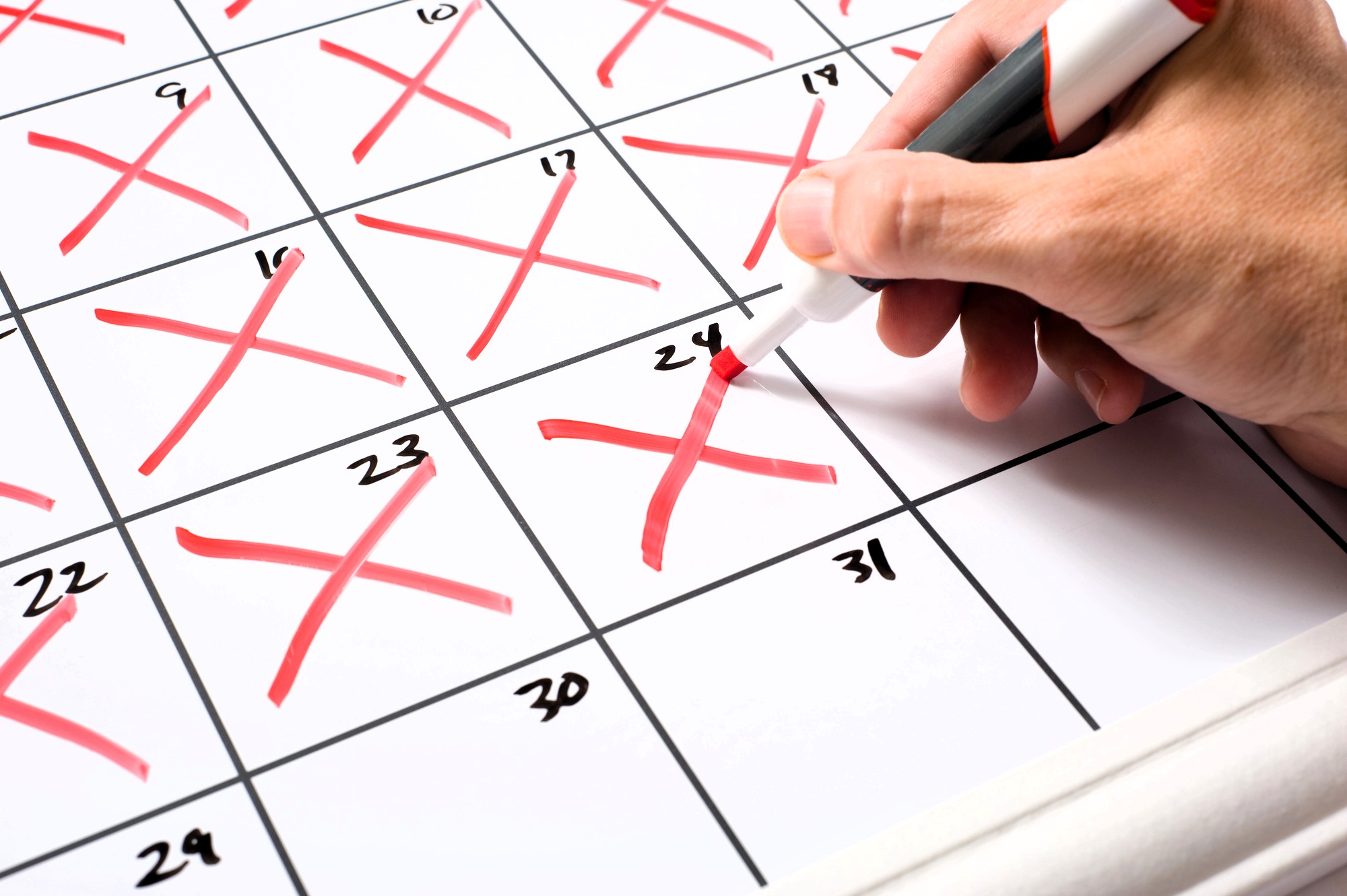 A hand holding a red marker crosses off the 24th day on a calendar. The calendar shows several days already marked with red Xs. The focus is on the hand and the calendar.
