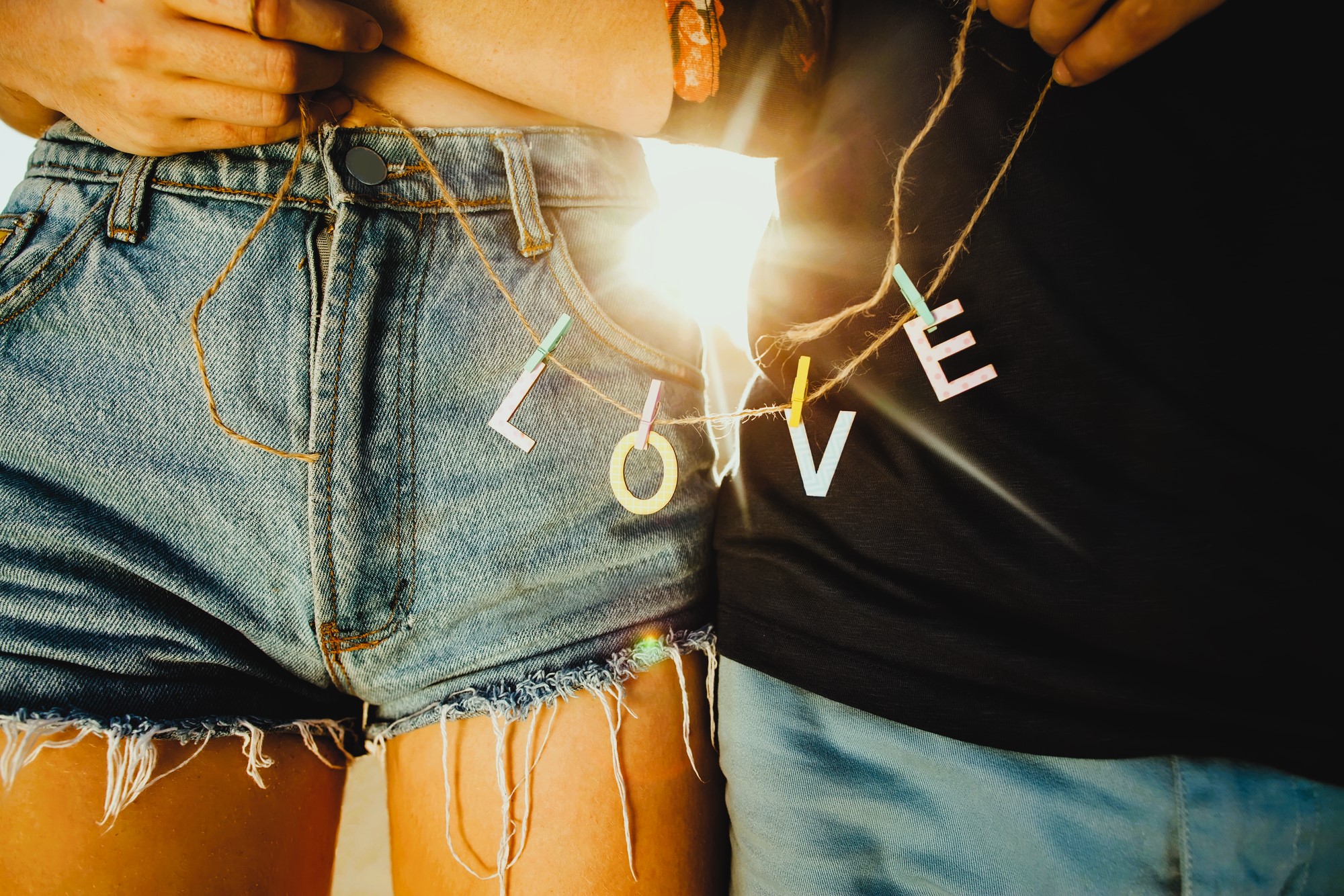 Two people stand close together, each holding a string with colorful letters spelling "LOVE" between them. They are wearing denim shorts and a black shirt, with sunlight shining through the letters. The mood is warm and intimate.