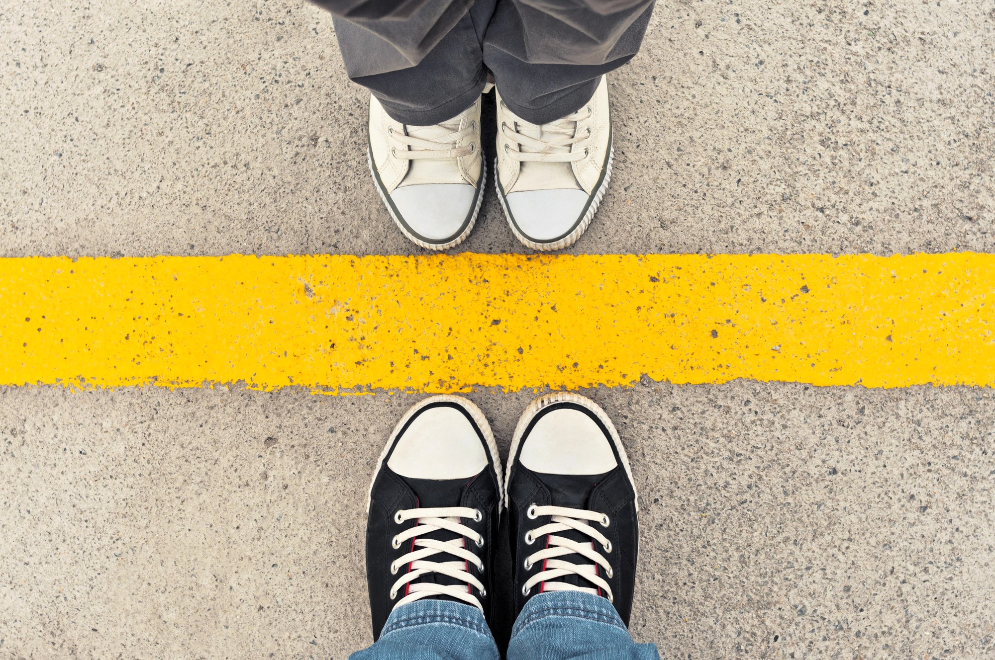 Two pairs of sneakers face each other on a concrete surface, separated by a bright yellow line. The top pair is white, and the bottom pair is black with white accents. The line creates a striking contrast against the grey background.