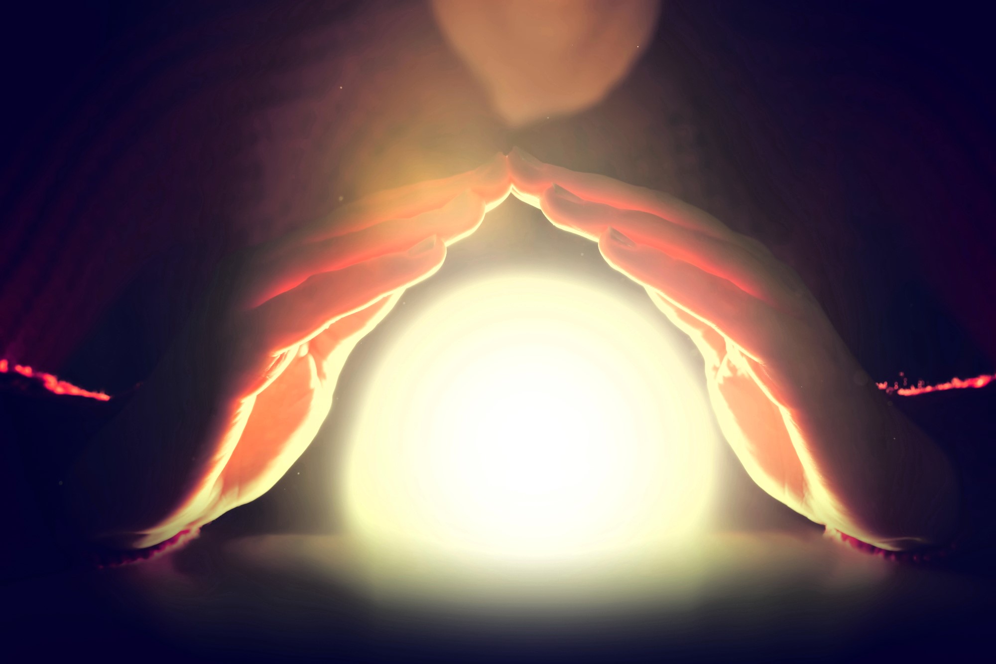 A pair of hands hover around a glowing, bright orb in a dimly lit setting, casting a mystical ambiance. The orb emits a warm light, illuminating part of the hands and the surrounding darkness.