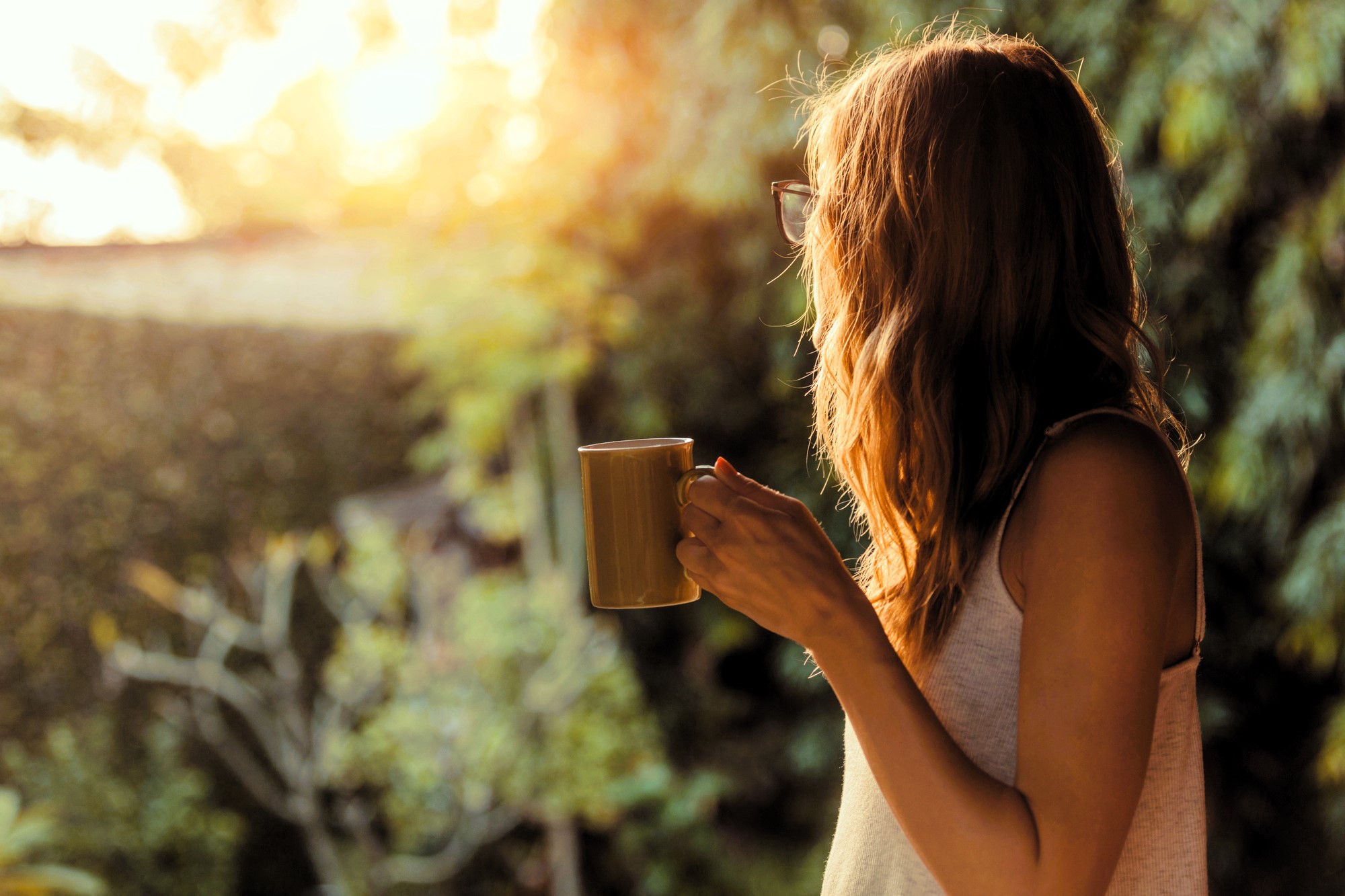 A person with long hair holding a coffee mug stands outdoors, gazing at a bright, sunlit scene surrounded by greenery. The soft sunlight creates a warm, golden glow.