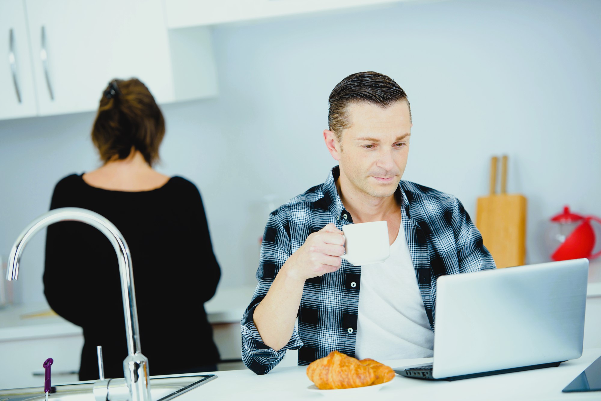 A man sits at a kitchen counter with a laptop and a croissant, holding a cup. He wears a plaid shirt. A woman in a black top stands with her back turned, near the kitchen sink. The setting is a modern, bright kitchen.