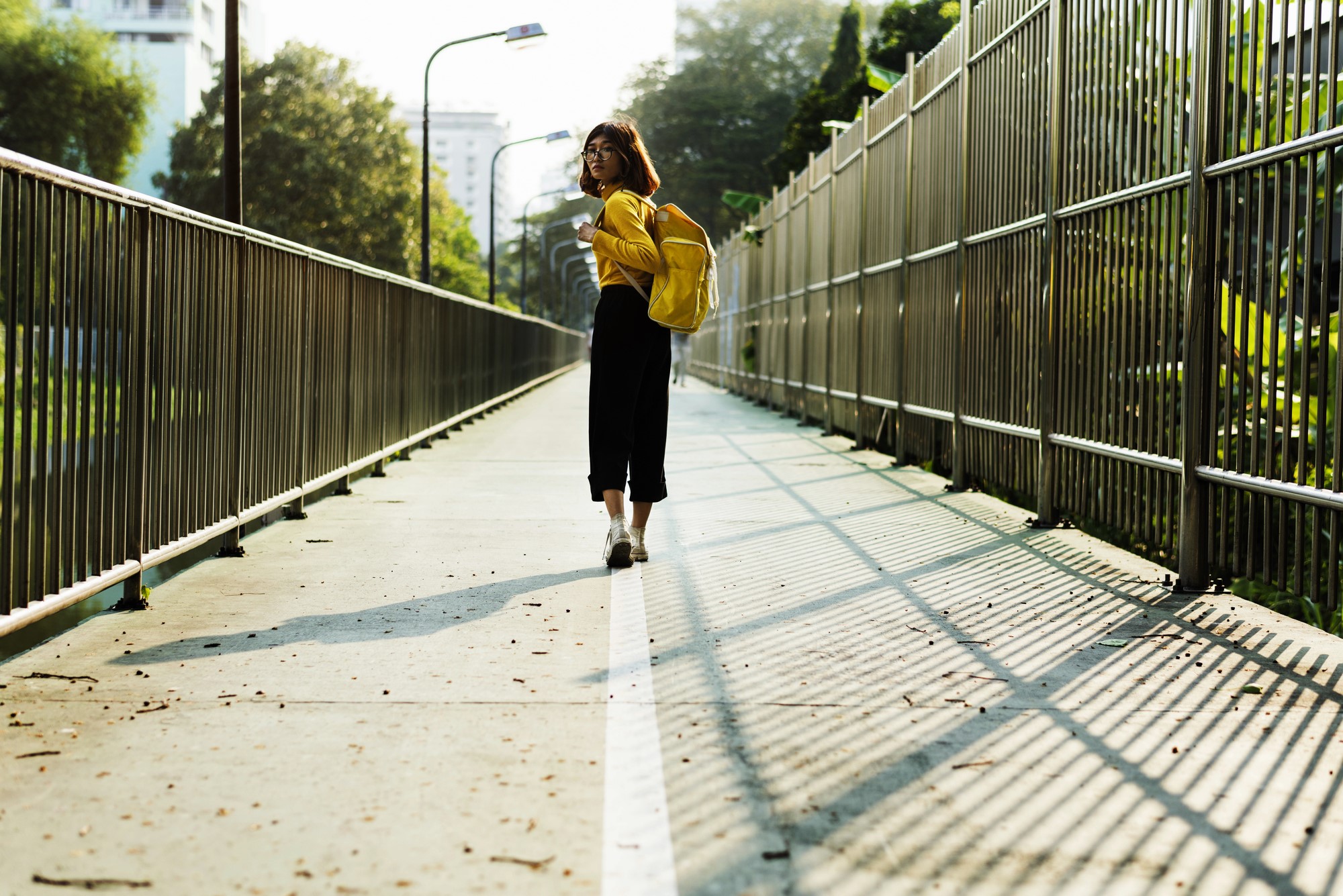 A woman wearing a yellow jacket and backpack walks on a pedestrian bridge surrounded by metal railings. The sun casts shadows of the railings on the walkway. Trees and buildings are visible in the background.