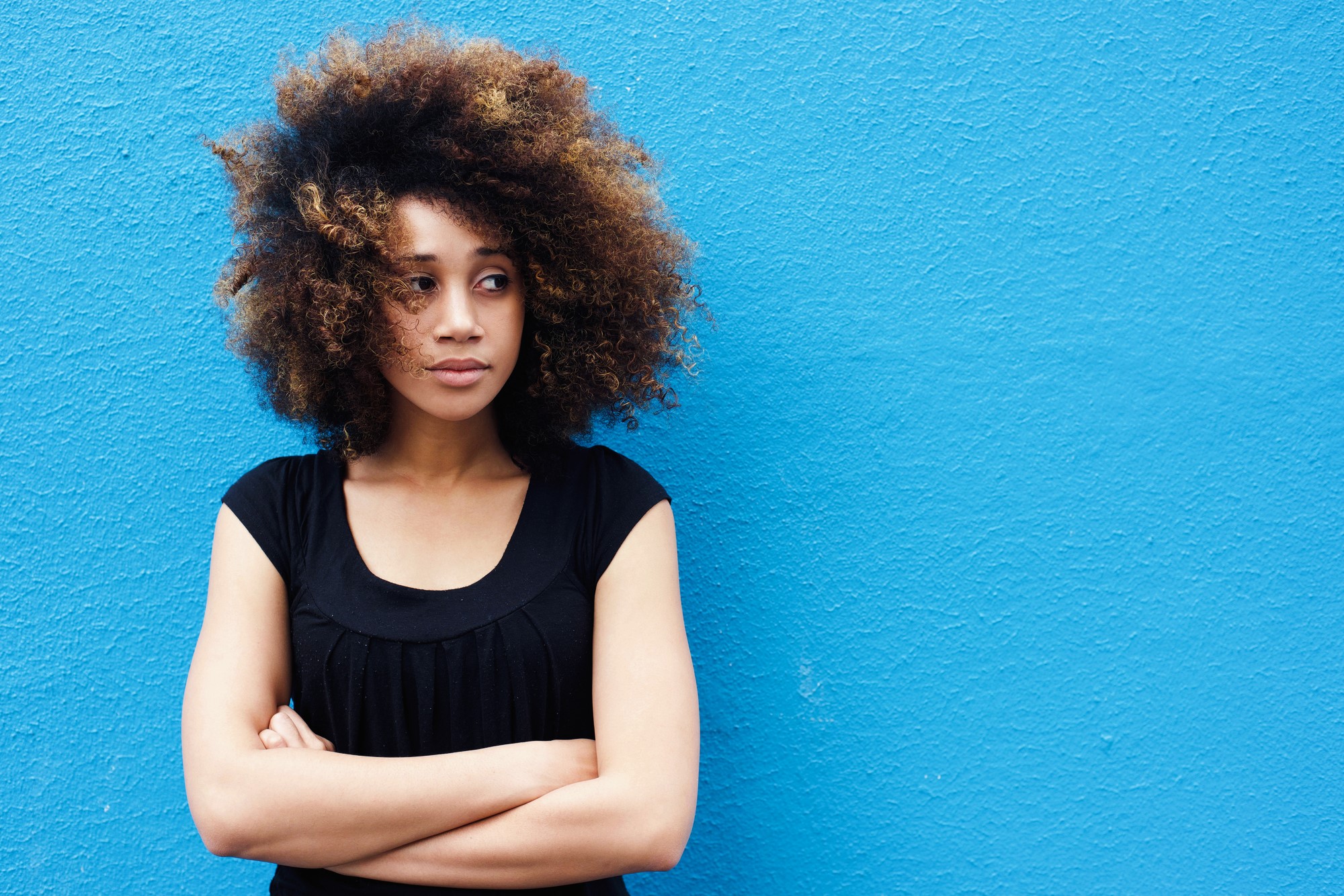 Person with curly hair standing against a bright blue wall, arms crossed and looking to the side. Wearing a black top.
