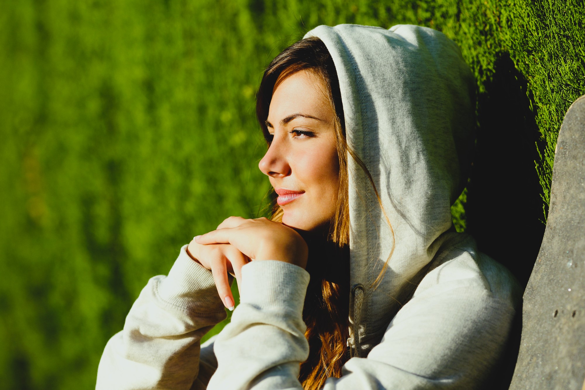Woman in a gray hoodie leans against a wall, gazing thoughtfully to the side with a serene expression. Sunlight highlights her features, and the background is a lush green hedge.