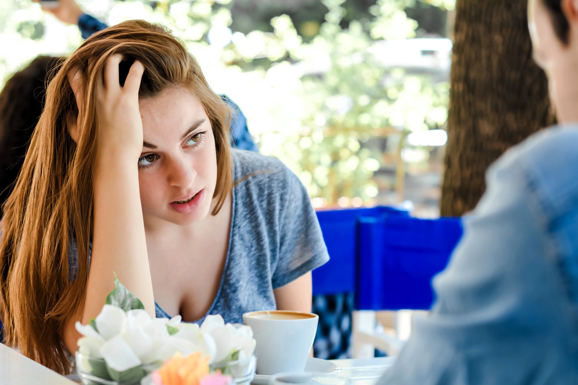 A woman with long brown hair, wearing a gray shirt, sits at a table, resting her head on her hand. She looks towards a person across from her. In front of her is a cup of coffee and a floral centerpiece. The background is blurred.