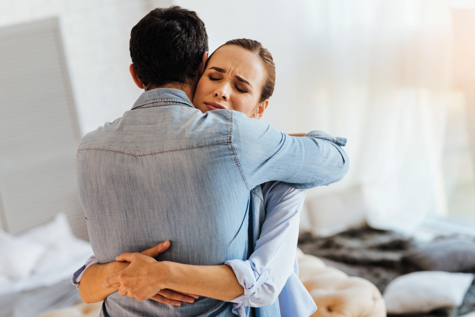 A woman and a man embrace in a comforting hug. The woman closes her eyes, appearing relieved or emotional. They stand in a softly lit room with a blurred background that includes a bed and a cozy ambiance.
