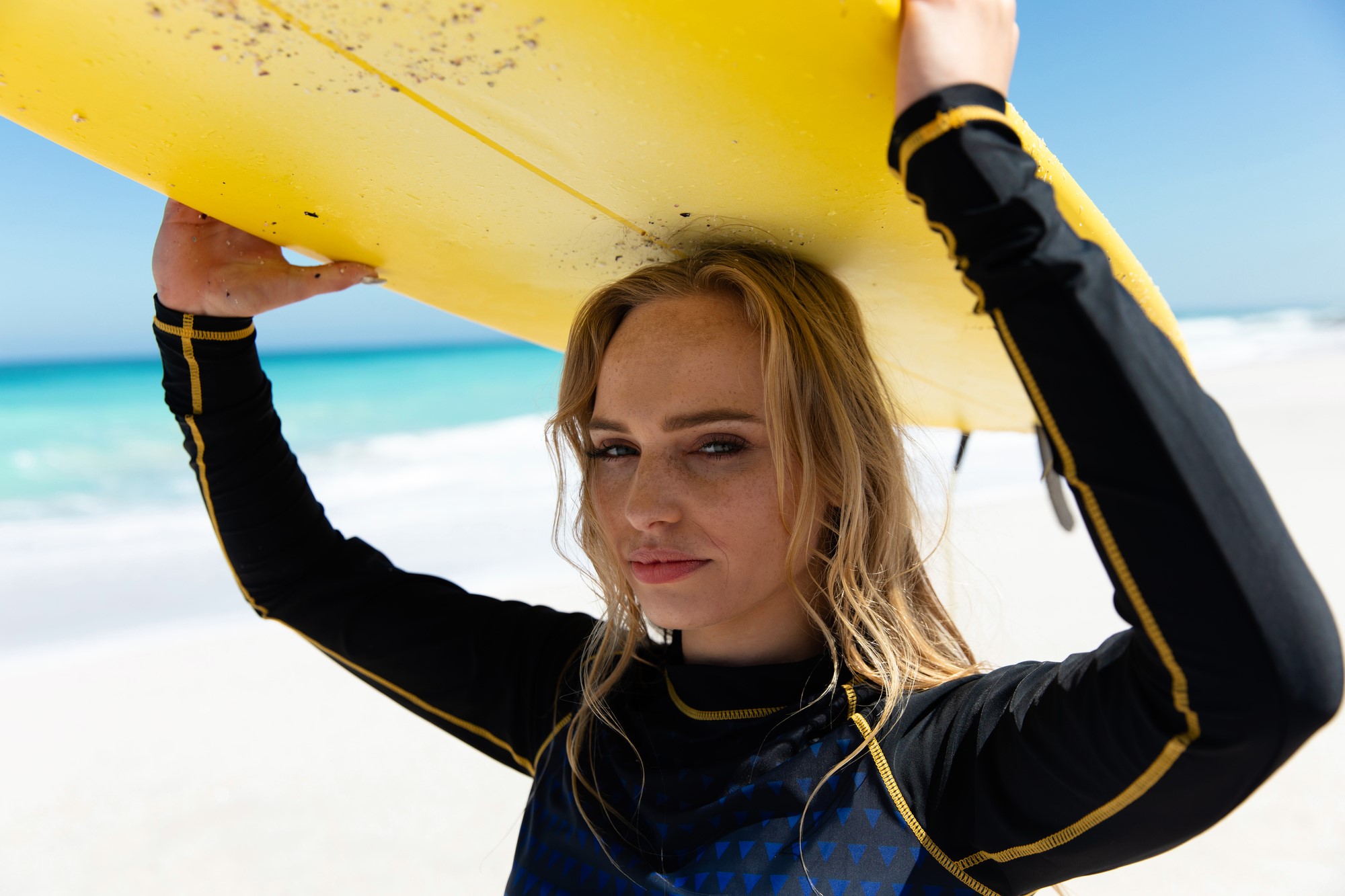 A woman on a beach holds a yellow surfboard above her head. She's wearing a black wetsuit with yellow accents, and the ocean is visible in the background under a clear blue sky.