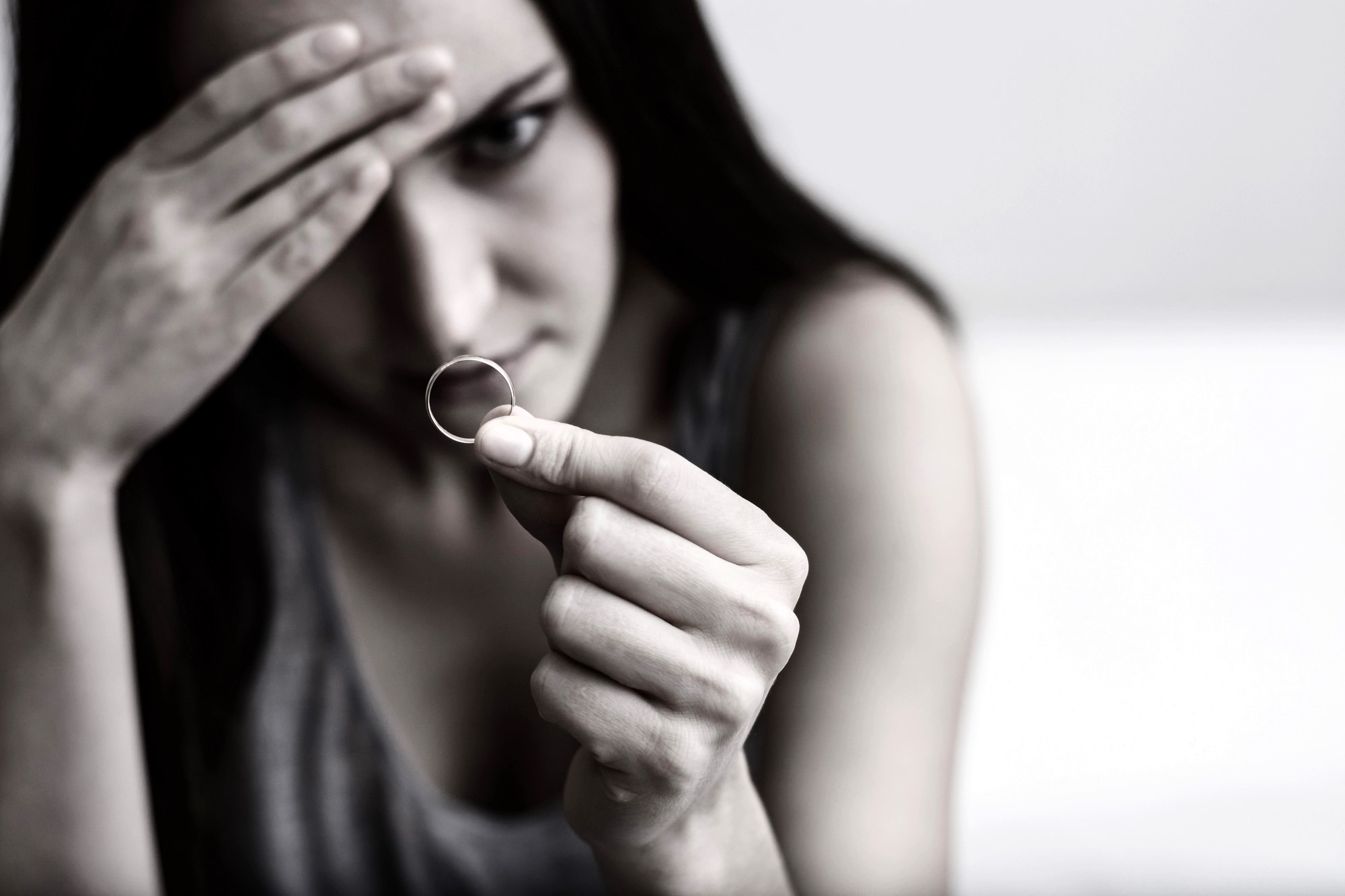 A woman with a pensive expression holds a ring in her hand. Her other hand rests on her forehead, partially obscuring her face. The image is in black and white, suggesting a somber or reflective mood.