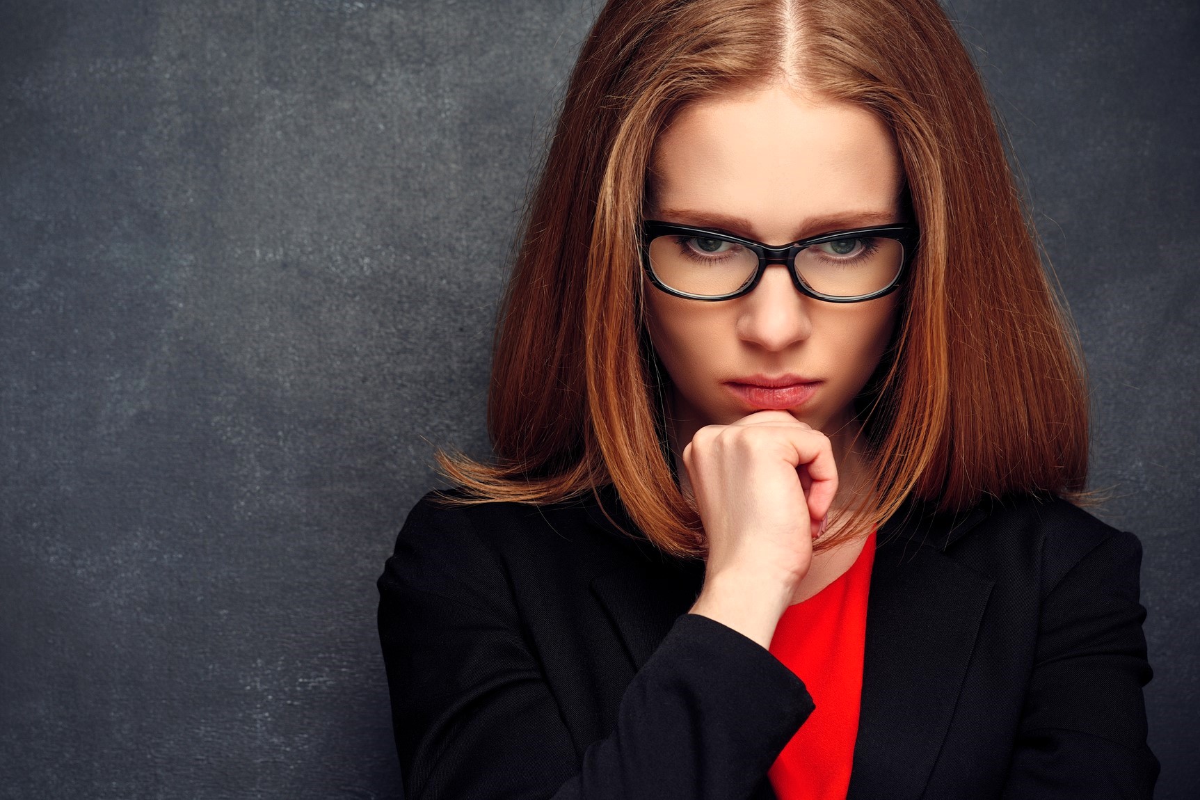 A woman with long auburn hair and glasses looks down pensively, resting her chin on her hand. She is wearing a black blazer over a red top, with a dark, textured background behind her.