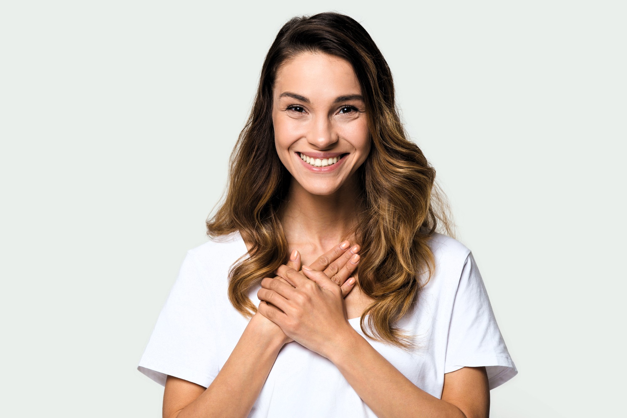 A person with long brown hair smiles warmly, hands clasped together at the chest. They wear a white T-shirt against a light grey background.