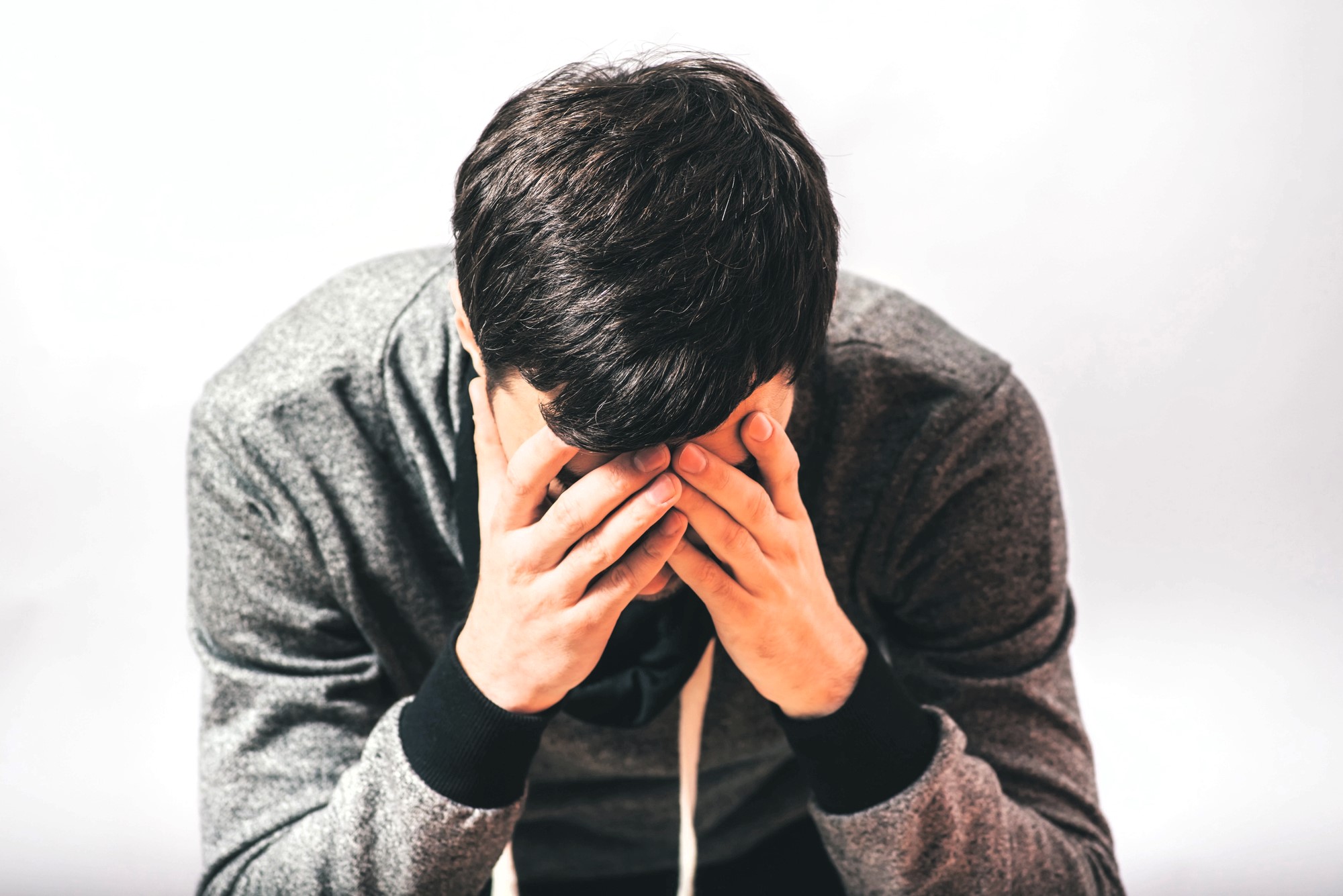 A person with short dark hair, wearing a gray hoodie, sits with their hands covering their face in a gesture of distress or contemplation against a plain background.